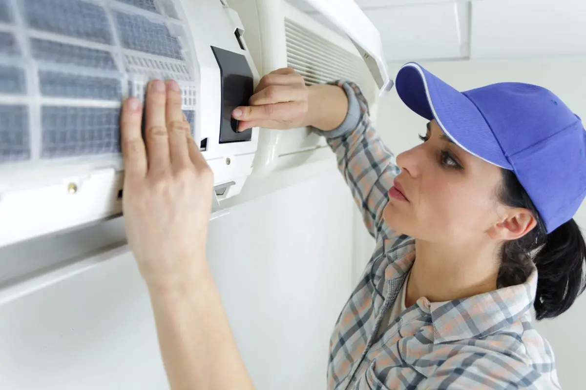 Image of a woman wearing a blue baseball cap working on an air conditioning unit. Source: adobe stock
