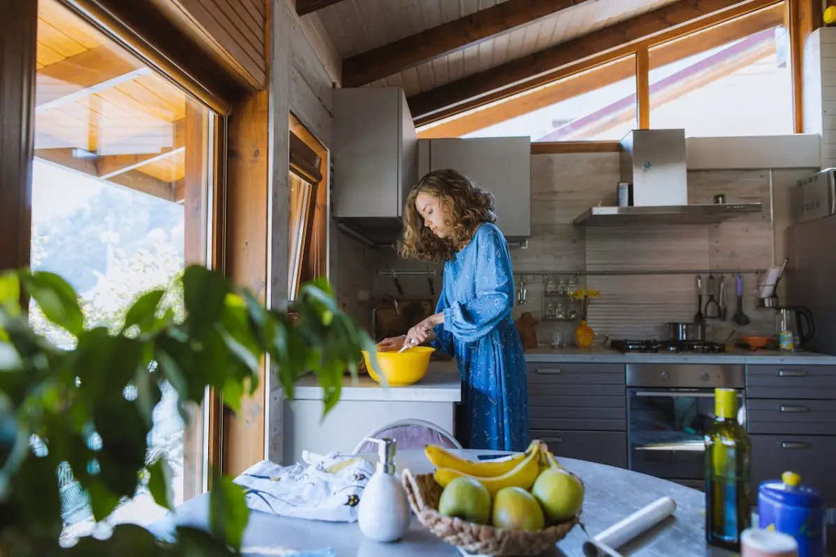 Image of a kitchen with a woman standing, while mixing near the countertop, cabinets hanging on the wall, and a range hood over a stove. Source: anastasia shuraeva, pexels